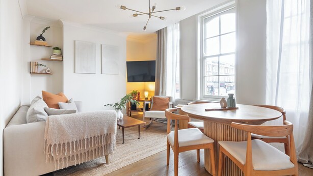 A modern living room with a sofa, a coffee table, and a dining table with chairs. There is a large window with curtains, a ceiling light fixture, and wall-mounted shelves with decorative items. - Centrally located Scandinavian high quality design. 2 min walk to station (London)