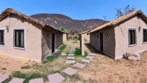 A row of rustic, single-story buildings with thatched roofs and a stone pathway leading through them. - Chokhiwadi Forest Retreat (Thanagazi)