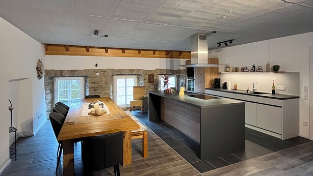 A spacious kitchen with a central island, a dining area with a wooden table and chairs, and a stone accent wall. - Gemütliches Ferienhaus für Große Familien und Gruppen (Kell am See)