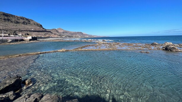 A coastal landscape with clear water, rocky shore, and distant mountains.