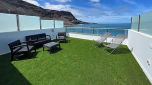 A rooftop terrace with artificial grass, seating, and a view of the ocean and cliffs.