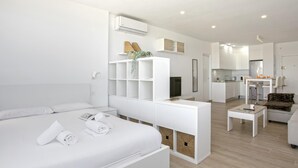 Una habitación de hotel moderna con cama, una mesita consola blanca, una pequeña zona de estar y una kitchenette.