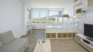 Una sala de estar moderna con sofá, mesa de centro, televisor y escritorio con silla. Amplias ventanas ofrecen vista a un paisaje montañoso.