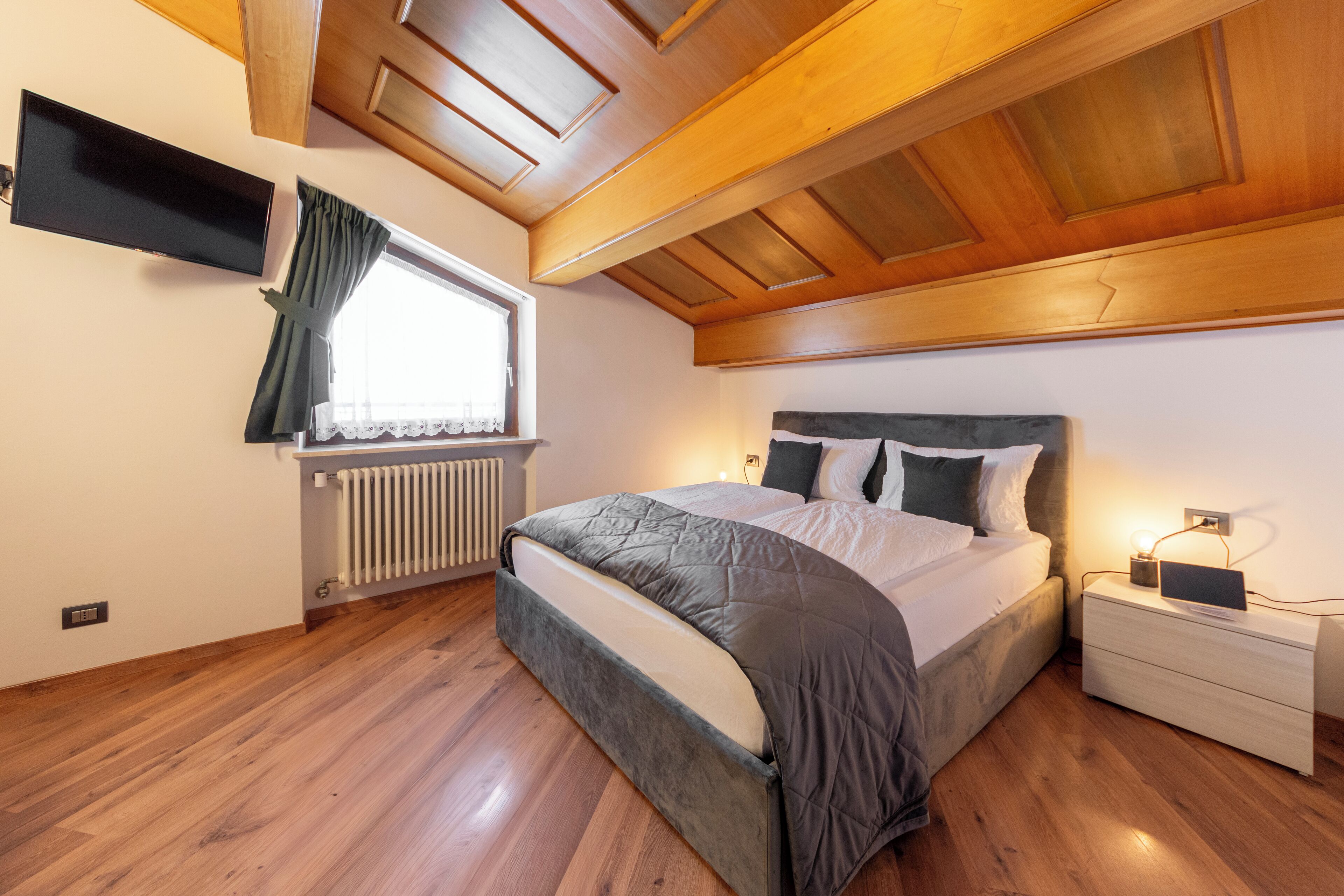 Une chambre à coucher comprenant un lit, une table de chevet, une fenêtre avec des rideaux, un radiateur et des poutres en bois au plafond.