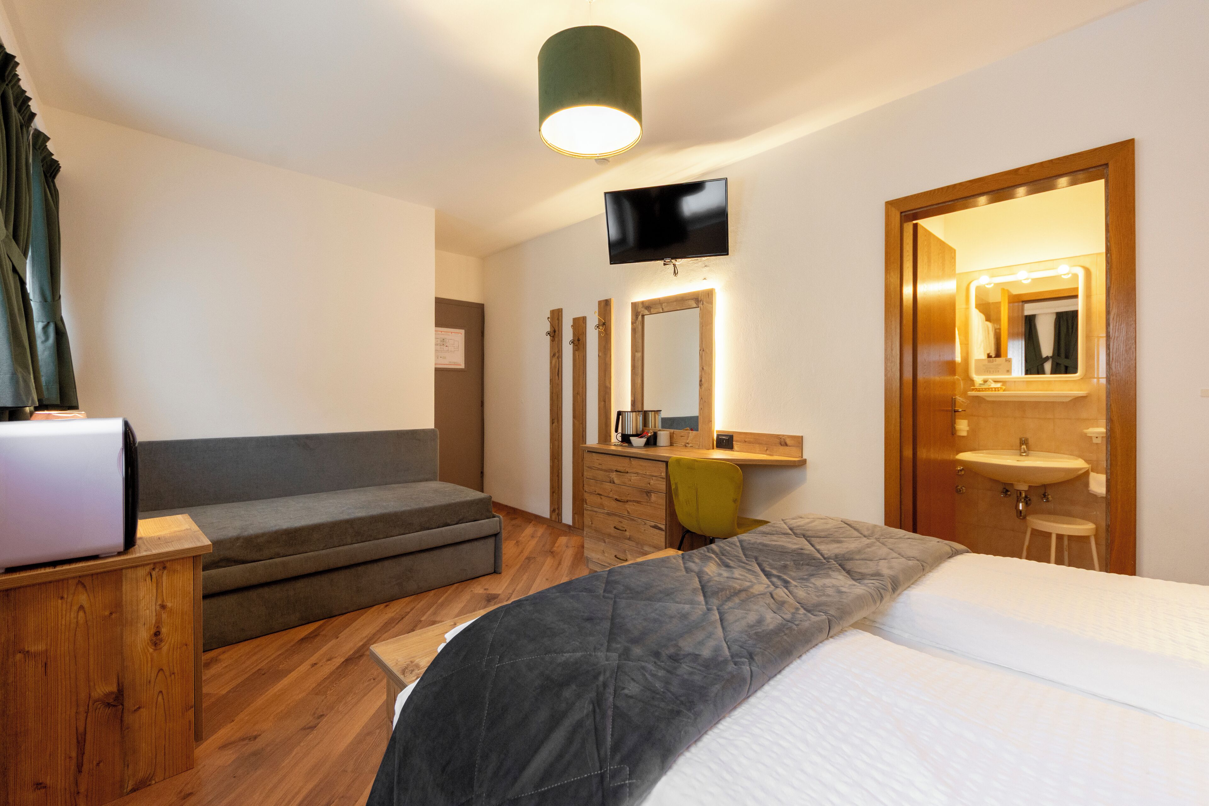 Hotellihuone, jossa on sänky, työpöytä kannettavalla tietokoneella, puinen lipasto, peili, televisio sekä kylpyhuone, jossa on pesuallas ja peili.