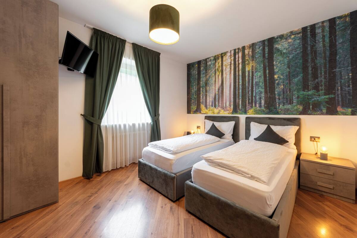 A hotel room with two beds, a TV mounted on the wall, a large forest-themed painting, and a wooden floor.