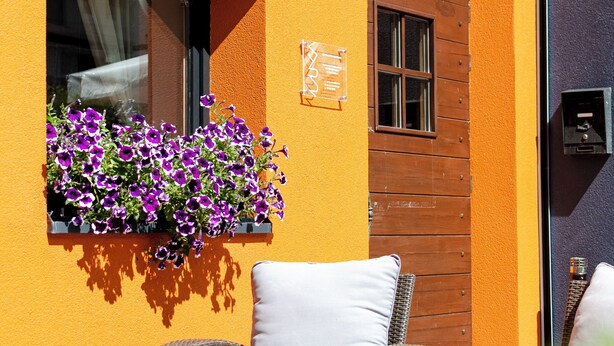 Ein Korbstuhl mit Kissen, ein Fenster mit violetten Blumen und eine Holztür.