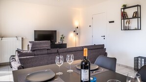 A modern living room with a dining area set for two. There is a bottle of wine on the table, two glasses, and a remote control on the sofa. - Artemis Gvo2 Apartment in Koksijde Near the Beach (Koksijde)