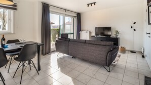 A modern living room with a grey sofa, a dining table with chairs, a TV, and a large window with curtains. - Artemis Gvo2 Apartment in Koksijde Near the Beach (Koksijde)