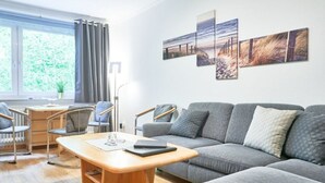 A modern living room with a grey sofa, a wooden coffee table, and a desk with a chair. There are three wall-mounted artworks depicting landscapes. - Juchmann Modern Retreat (Timmendorfer Strand)
