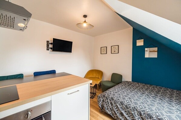 A compact bedroom with a bed, a desk with a chair, a TV mounted on the wall, and a ceiling light.