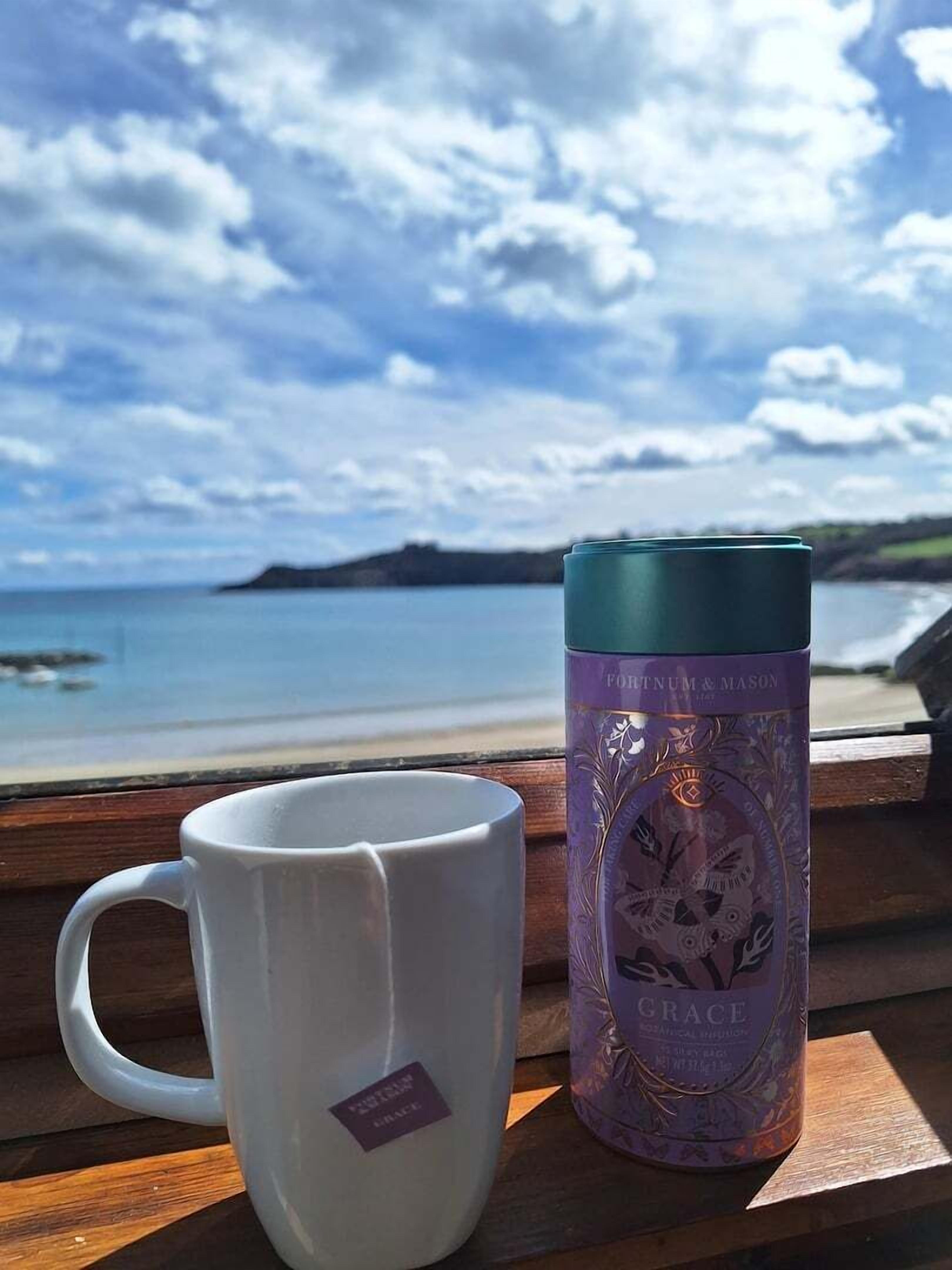 Una taza blanca y un termo púrpura sobre un banco de madera, con una vista panorámica de una playa y el océano al fondo.