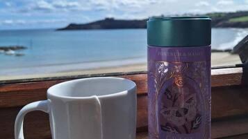 Una taza blanca y un termo púrpura sobre un banco de madera, con una vista panorámica de una playa y el océano al fondo.
