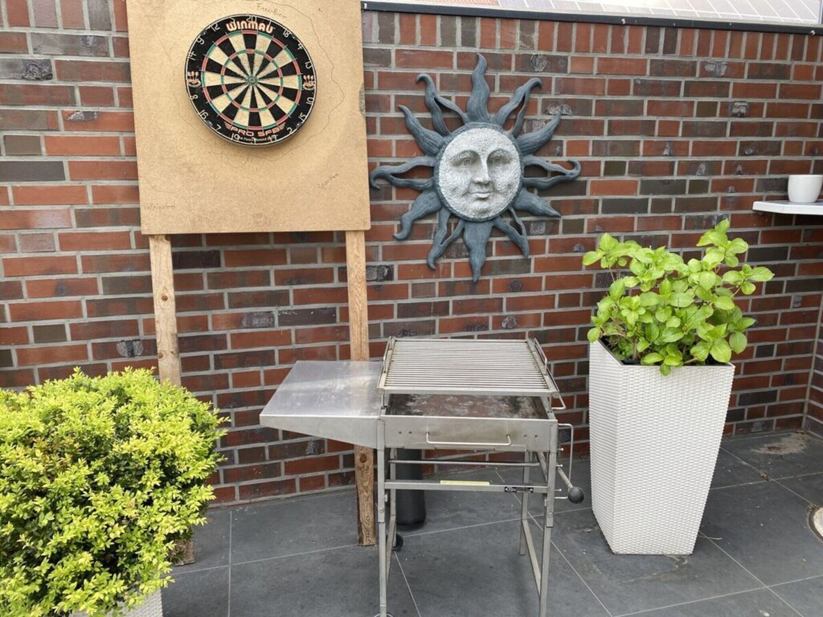 A dartboard, a sun-shaped wall decoration, a metal table, and a potted plant against a brick wall.