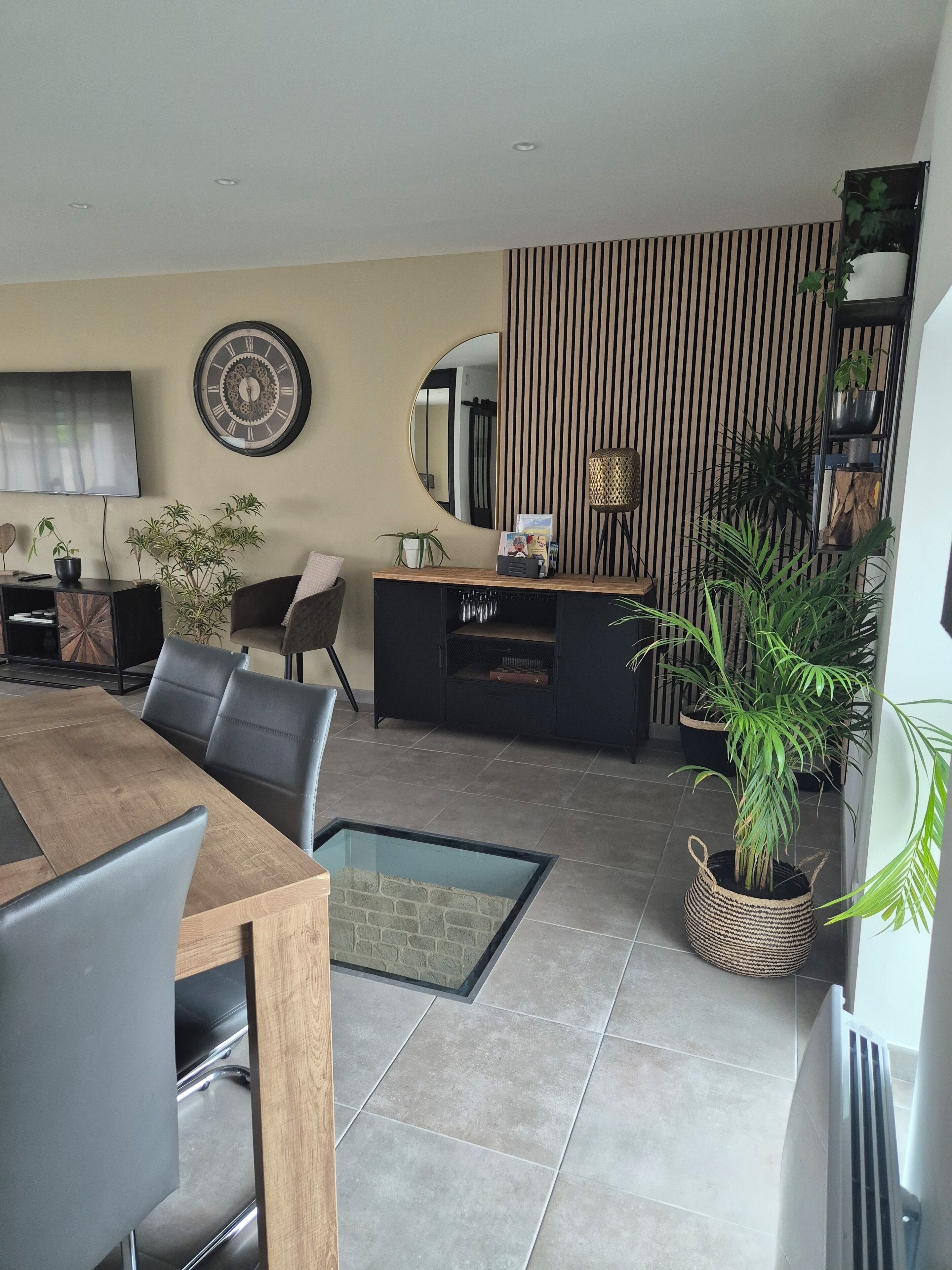 Un salon moderne avec une table à manger en bois, des chaises en cuir noir, un téléviseur et un grand miroir.
