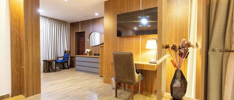 A hotel room with a wooden floor, a desk with a chair, a television mounted on the wall, and a vase with dried flowers on a shelf.