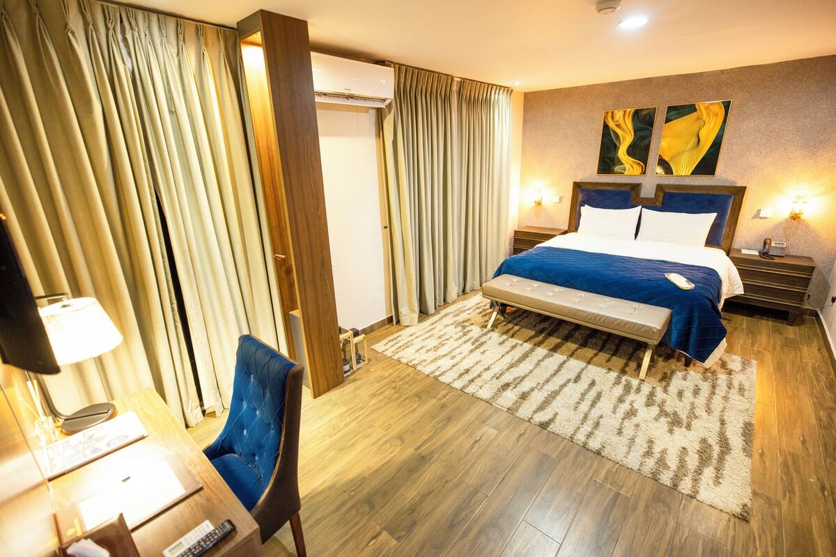 A hotel room with a bed, a desk with a lamp, a chair, and a large window with curtains.