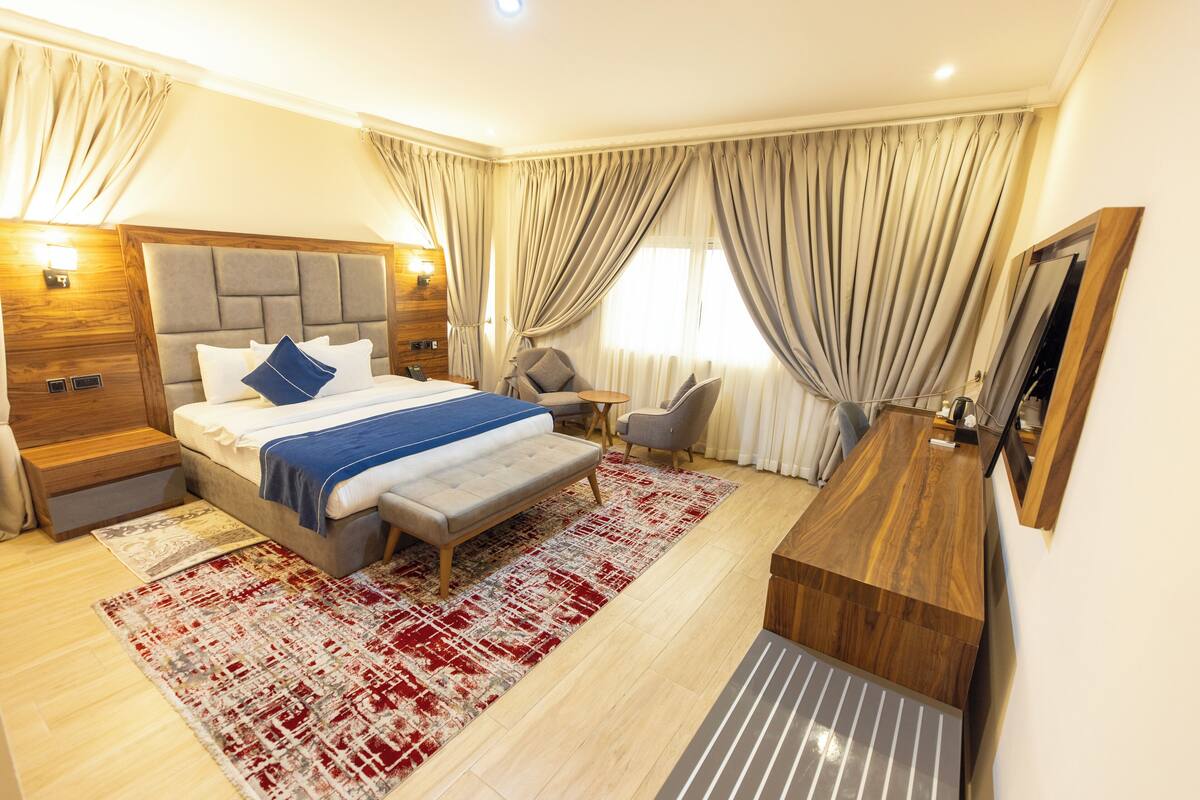 A modern hotel room with a large bed, a wooden desk, a chair, and a small table.