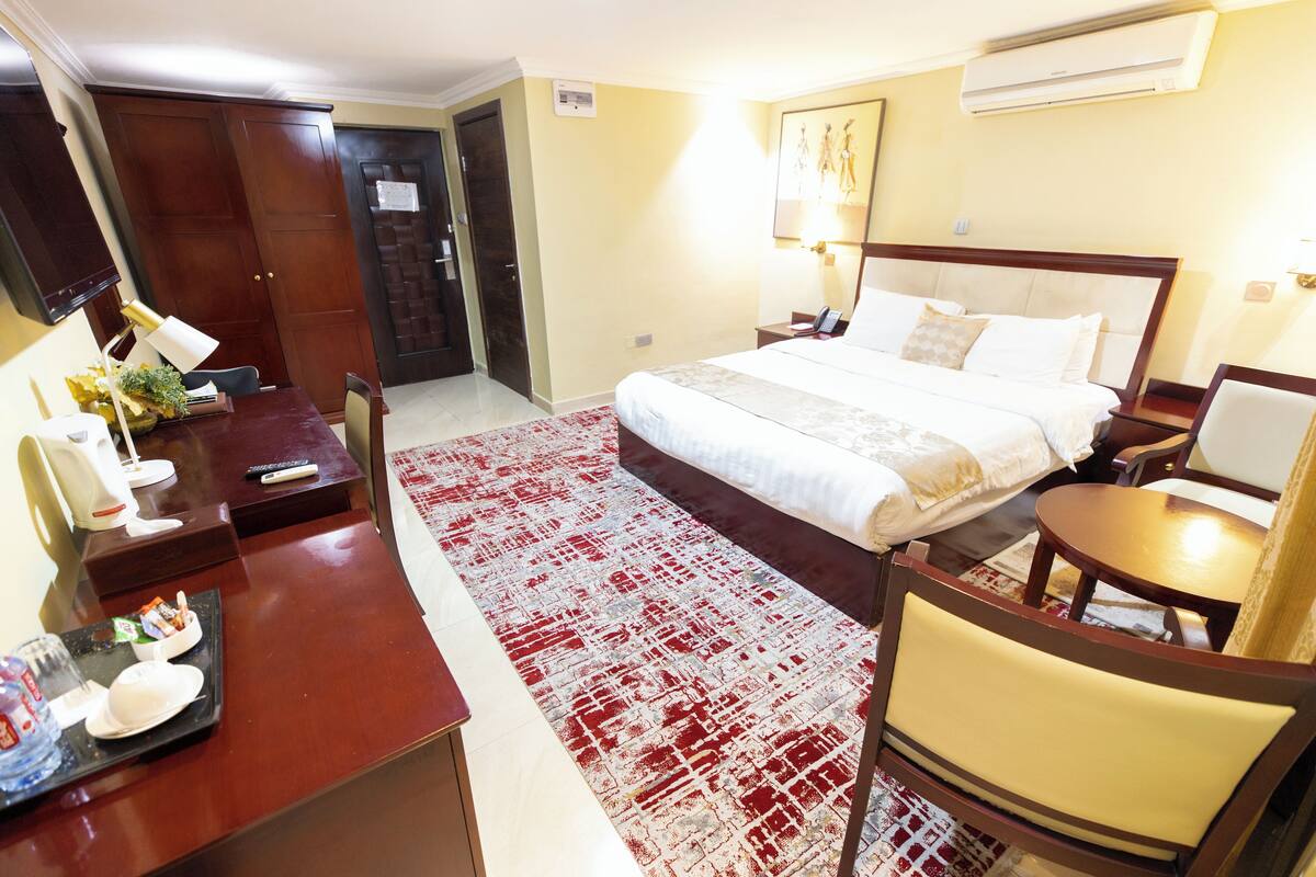 A hotel room with a bed, bedside table, chair, desk, and a red and white patterned rug.