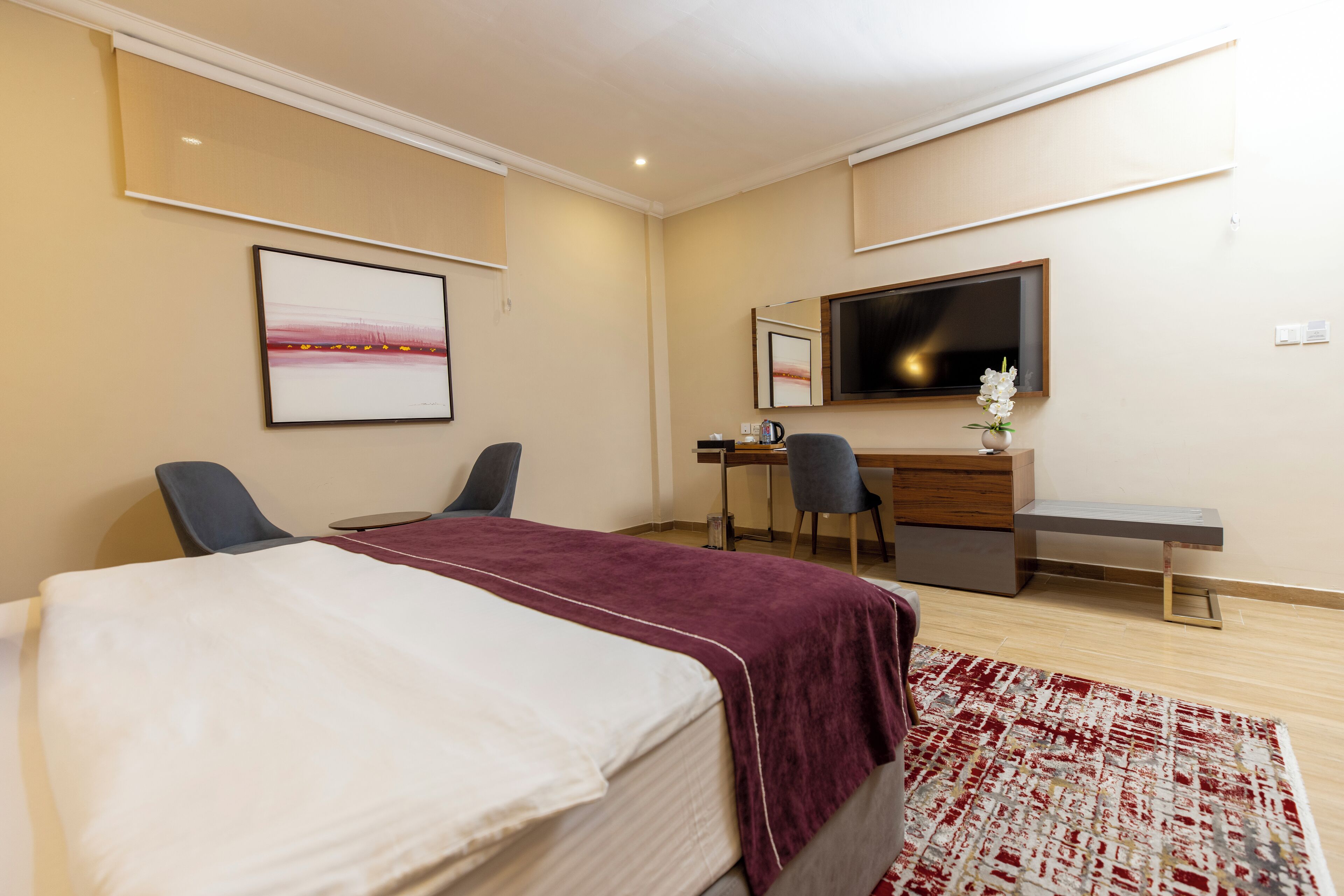 A hotel room with a bed, two chairs, a desk, a TV, and a small plant.
