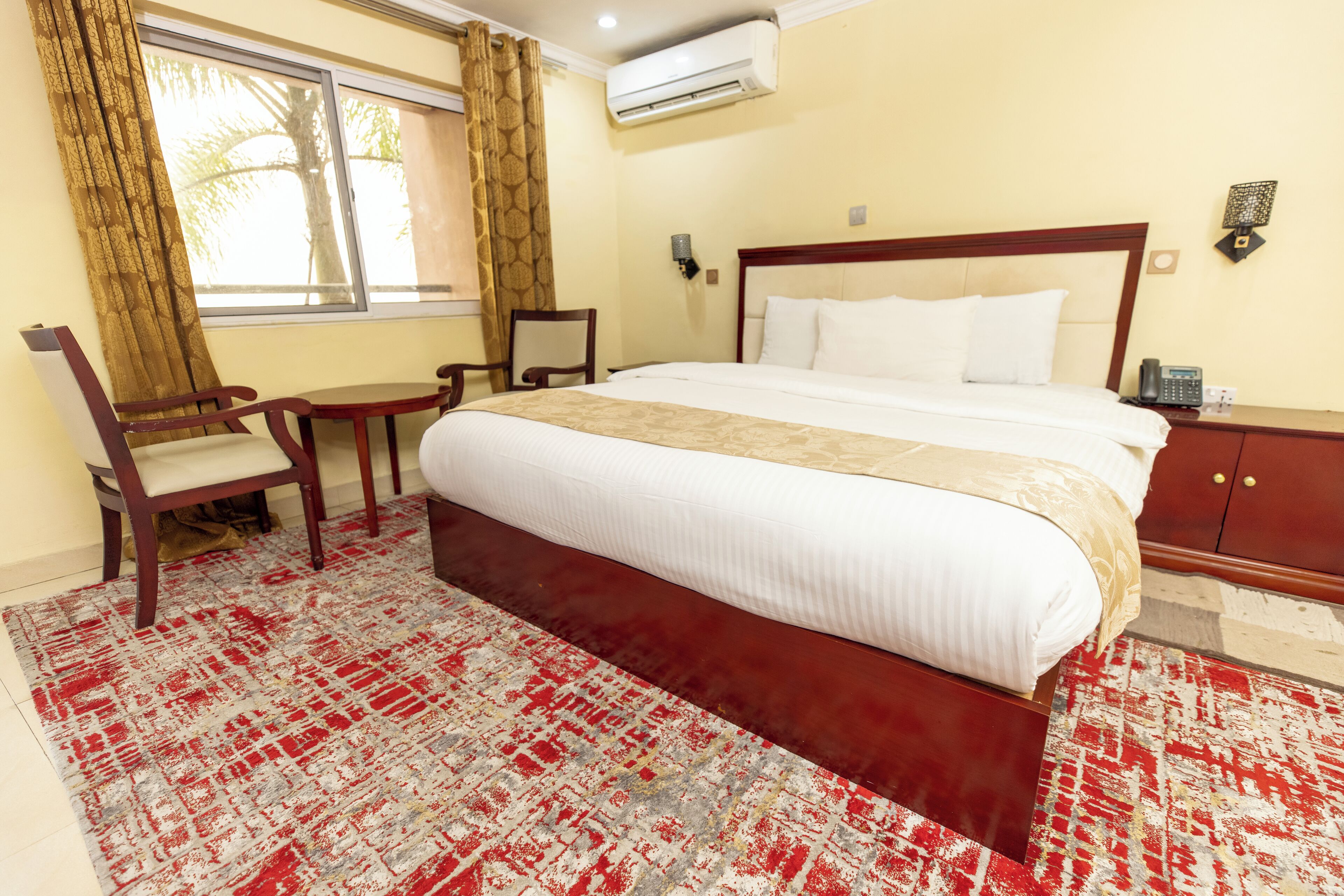 A hotel room with a large bed, a wooden nightstand, a desk with a chair, and a window with curtains.