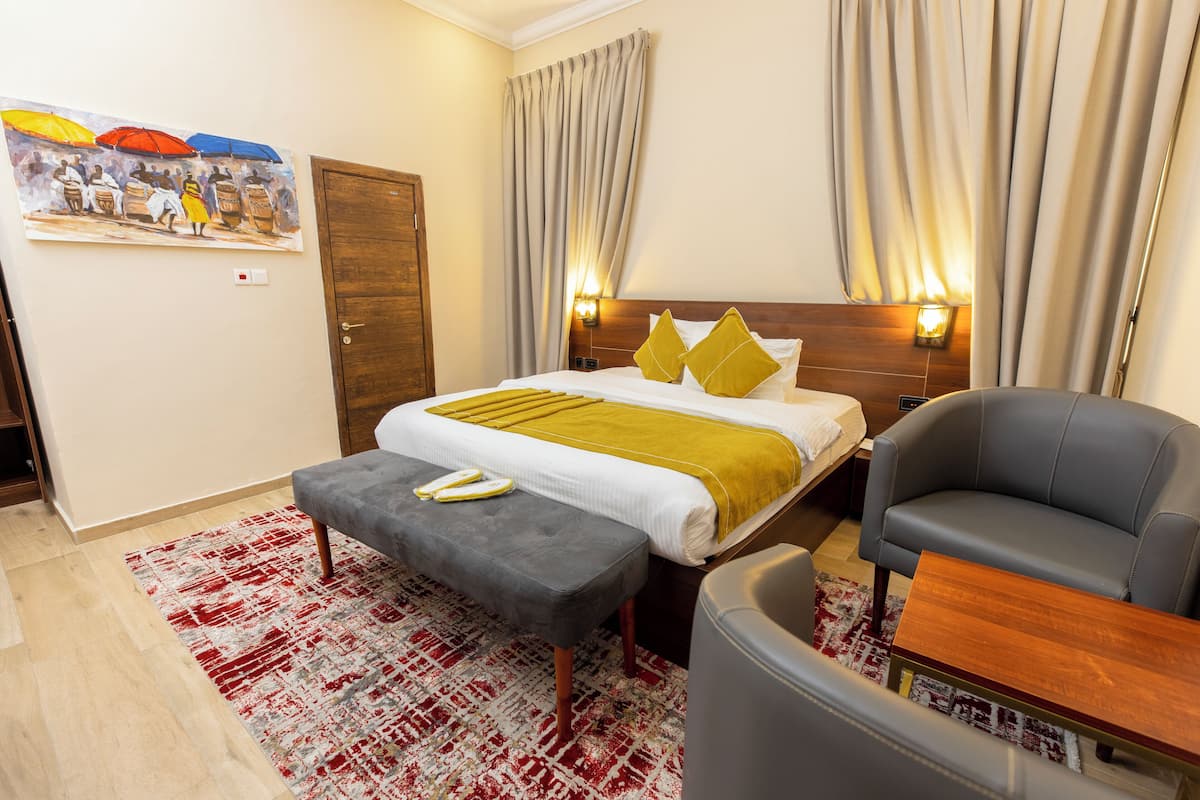 A hotel room with a bed, a grey armchair, a wooden side table, a red and white patterned rug, and a painting on the wall.