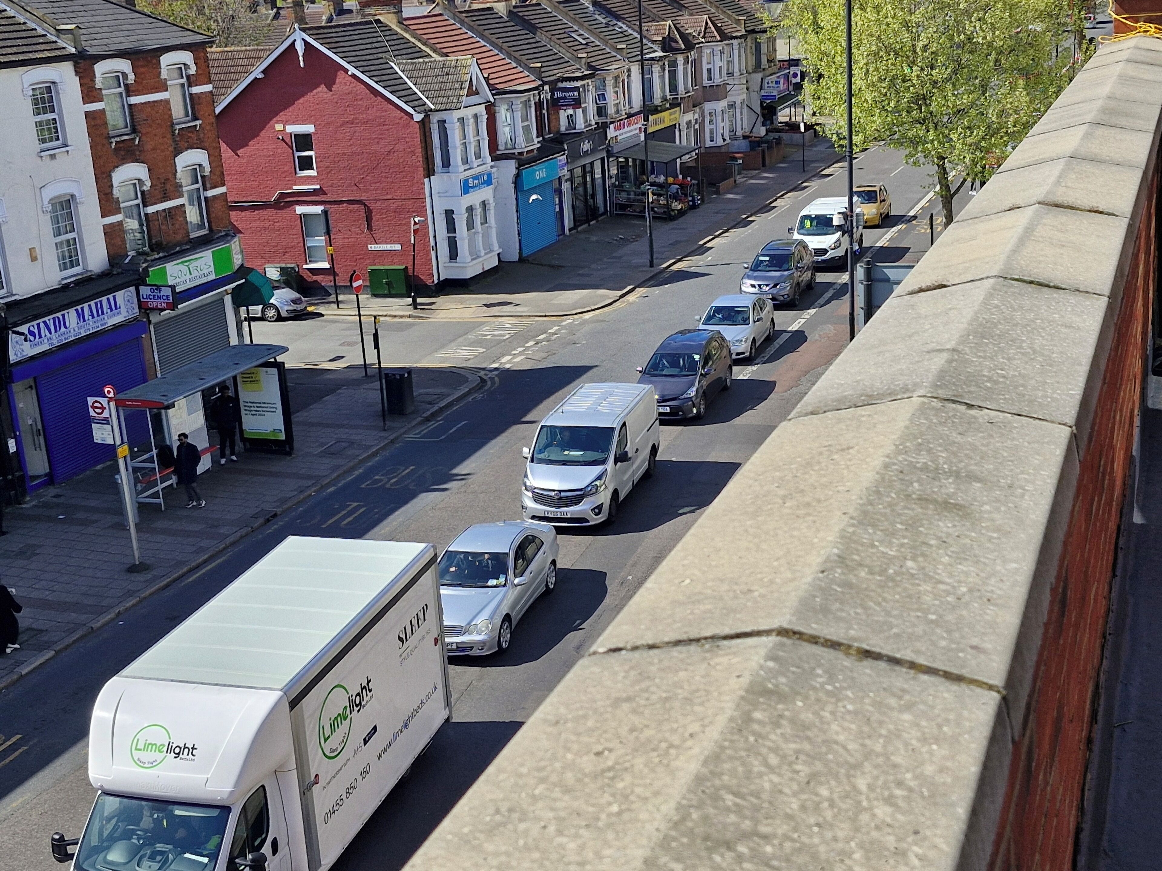 A street view with parked cars, a white delivery truck, and buildings with various shops.