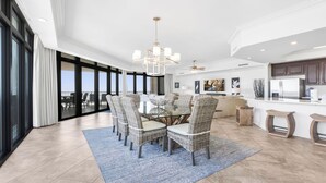 A spacious dining area with a glass-top table surrounded by wicker chairs, a modern kitchen with dark cabinets, and a view of the outdoors through large sliding doors. - Beachfront!  Lazy River, Restaurant, New condo! (Orange Beach)