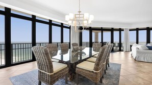 A spacious dining area with a glass table surrounded by wicker chairs, a chandelier, and a balcony with a view of the ocean. - Beachfront!  Lazy River, Restaurant, New condo! (Orange Beach)