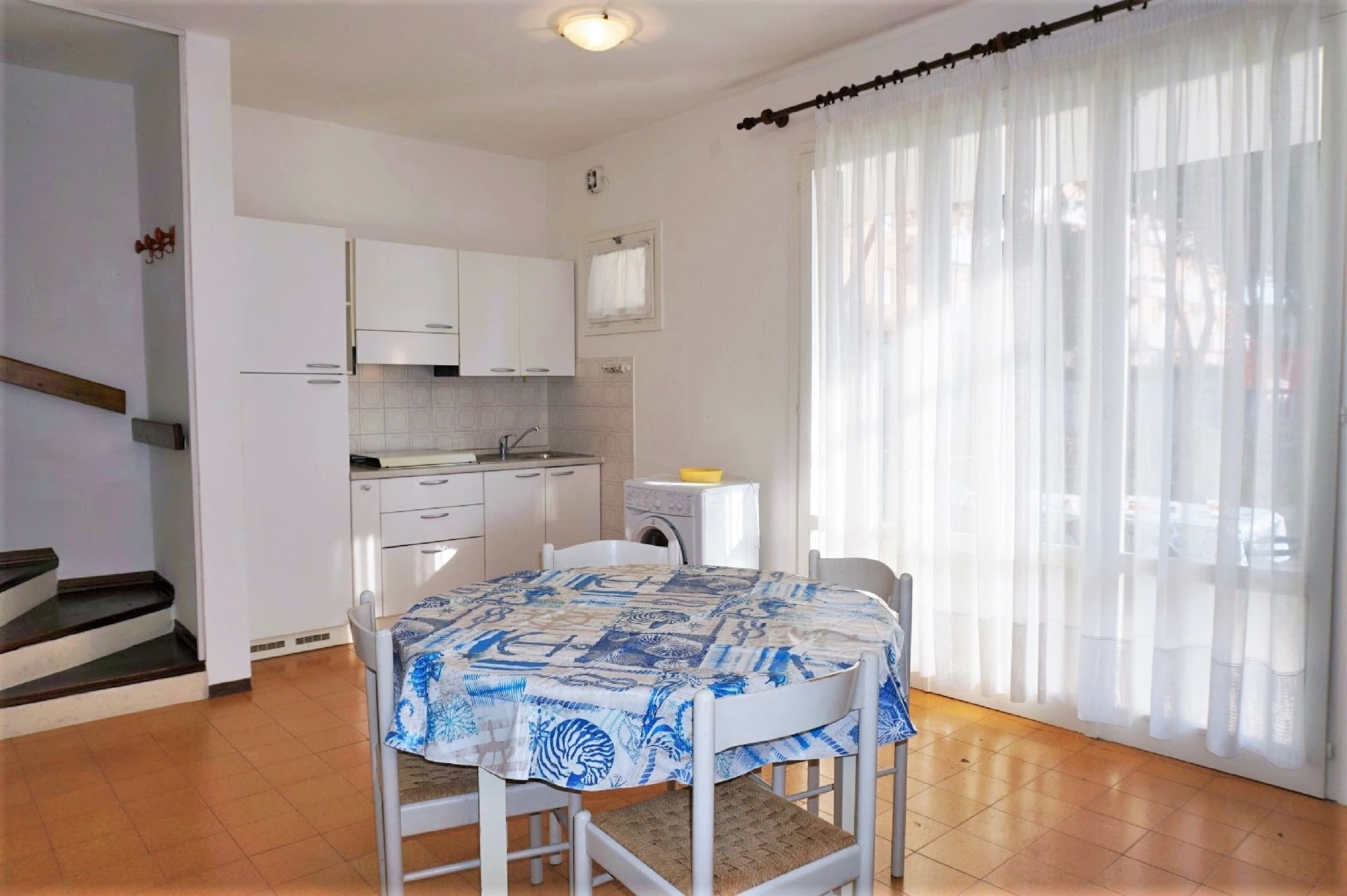 A kitchen with white cabinets, a dining table with a patterned tablecloth, and a window with sheer curtains.