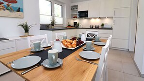 A modern kitchen with white cabinets, a wooden dining table set for breakfast, and a painting on the wall. - Villa Ostseekind Comfortable Holiday Residence (Ostseebad Dierhagen)