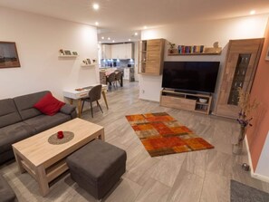 Una sala de estar moderna con un sofá, una mesa de centro y un televisor. Se observa una cocina al fondo con armarios blancos y un comedor.