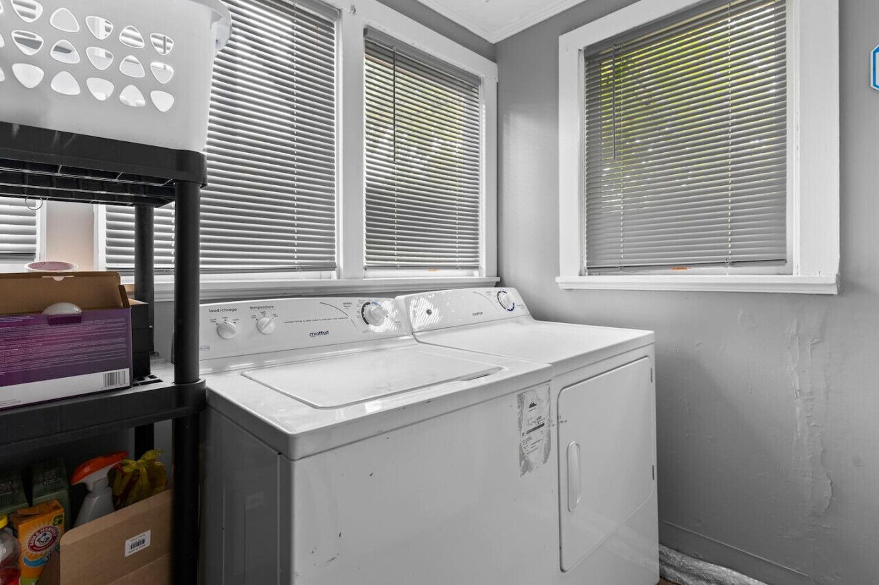 A laundry room with a front-loading washing machine, a shelf with cleaning supplies, and windows with blinds.