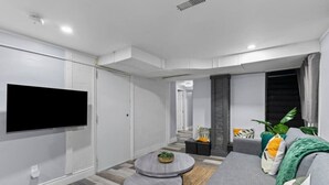 A modern living room with a flat-screen TV mounted on the wall, a grey sofa with cushions, a round coffee table, and a small side table with a plant.