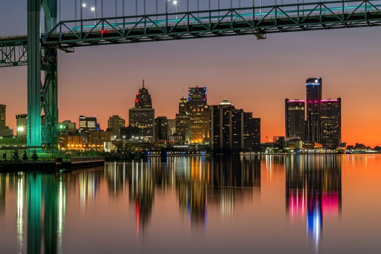 A nighttime cityscape with a prominent bridge and skyscrapers reflected in calm waters.