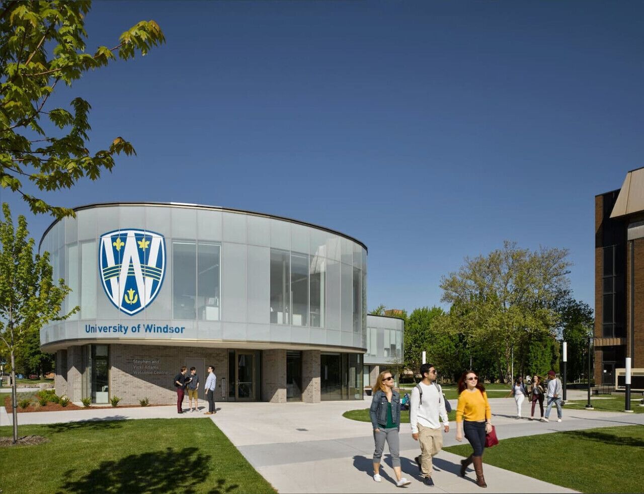 En moderne bygning med en glasfacade og et logo ved indgangen, omgivet af græsareal og et par personer, der går rundt.