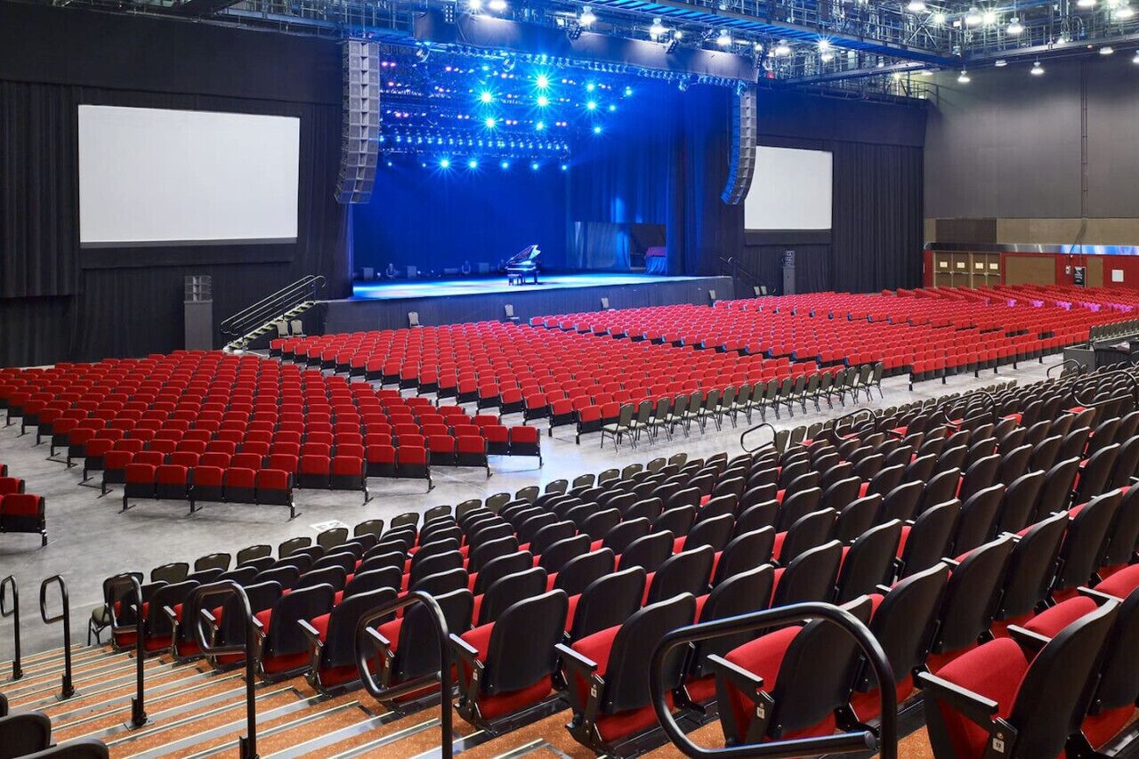 A large indoor auditorium with rows of red and black seating, a stage with a piano, and a large screen.