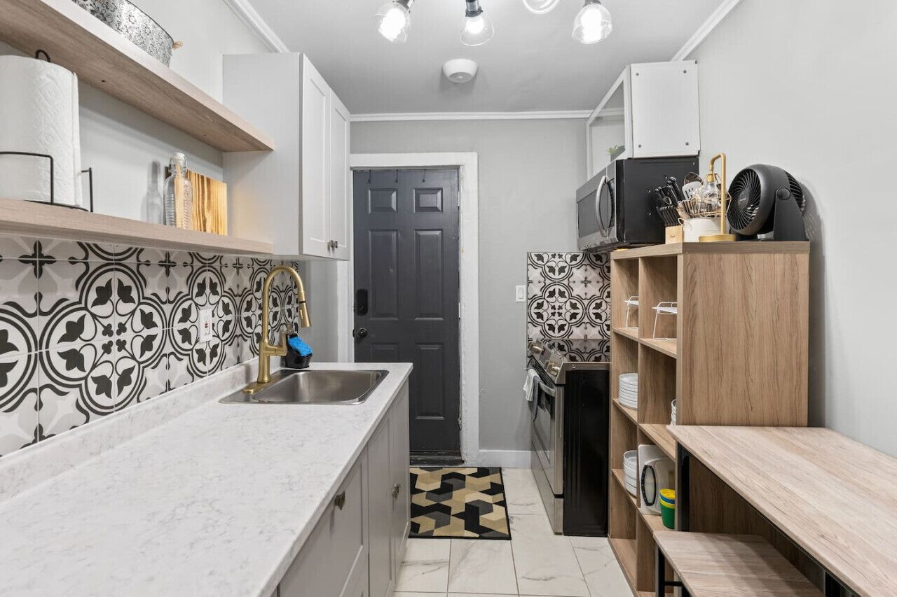 A narrow kitchen with a marble countertop, black and white patterned backsplash, and a black door in the background.