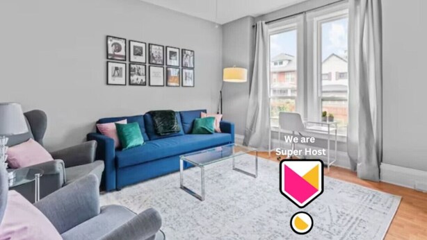 A modern living room with a blue sofa, grey armchairs, and a glass coffee table. There are framed pictures on the wall and a window with curtains. - Stylish 2bd  DT Laundry and Parking (Windsor)