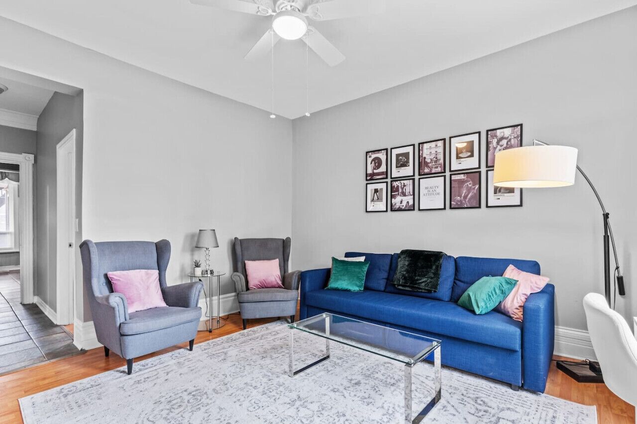 Un salon moderne avec un fauteuil gris, un canapé bleu et une table basse en verre. Des tableaux encadrés ornent le mur, et une lampe sur pied est placée dans un coin.