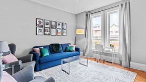 A modern living room with a blue sofa, grey armchairs, a glass coffee table, and a large window with curtains. - Stylish 2bd  DT Laundry and Parking (Windsor)