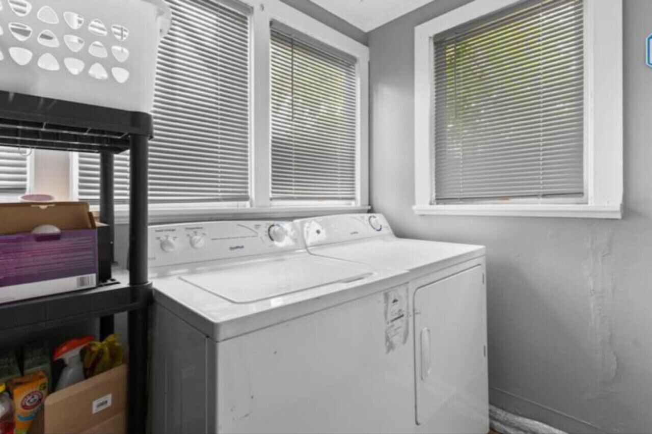 A laundry area with a front-loading washer and dryer, a shelf with cleaning supplies, and a window with blinds.