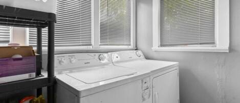 A laundry area with a front-loading washer and dryer, a shelf with cleaning supplies, and a window with blinds.