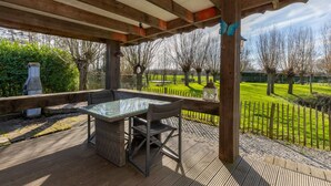 Une terrasse extérieure couverte, dotée d’une table et de chaises à plateau en verre, entourée d’une terrasse en bois et offrant une vue sur un champ verdoyant et des arbres.