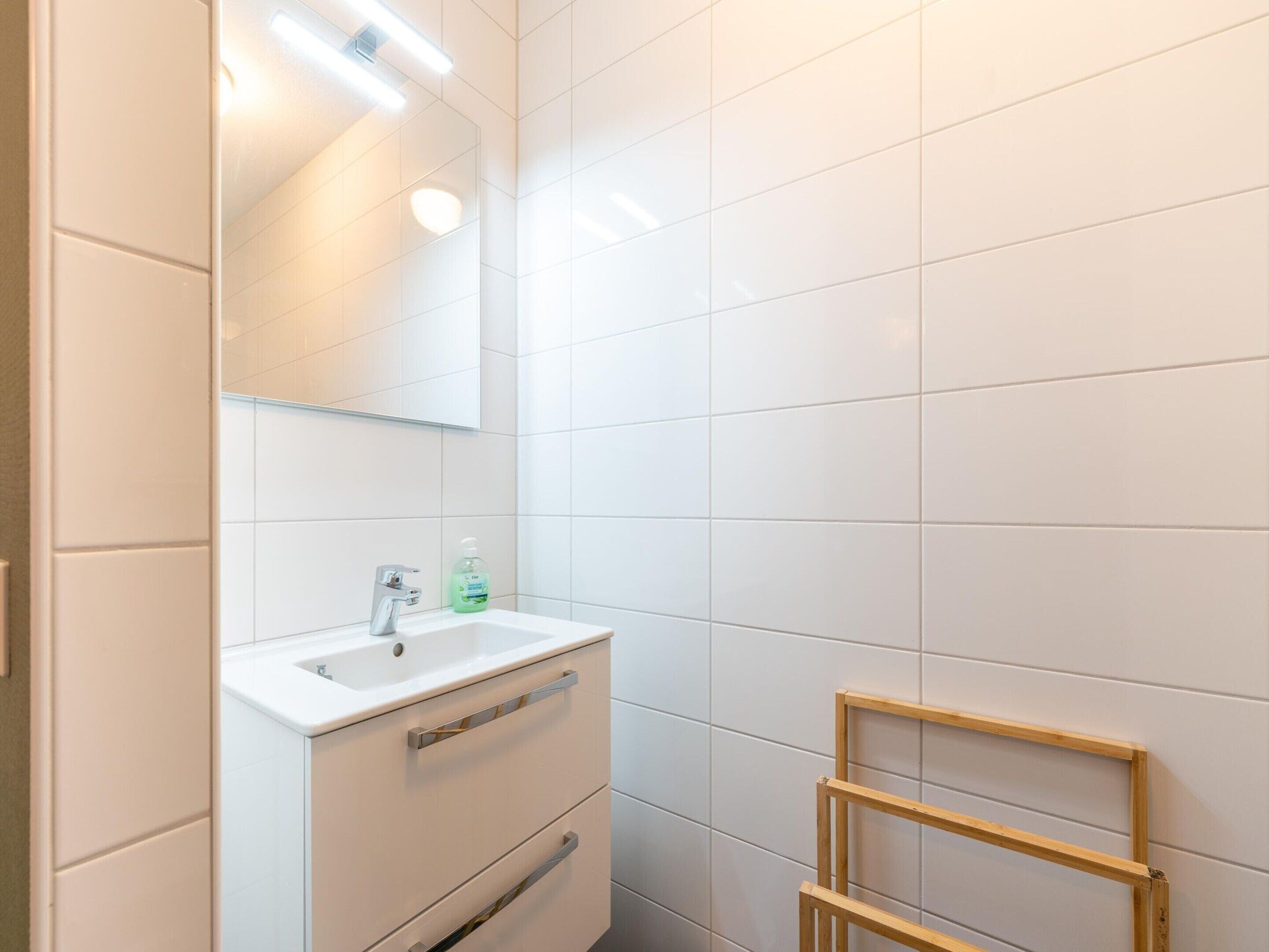 Un baño con azulejos blancos, un lavamanos blanco, un espejo y un estante de madera.