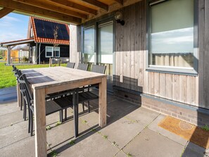An outdoor patio with a wooden table and chairs, a covered area, and a view of a house with a red roof and large windows.