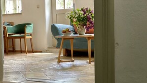 Een kamer met een stenen vloer, een groene bank, een houten tafel met een schaal fruit en een vaas met bloemen.