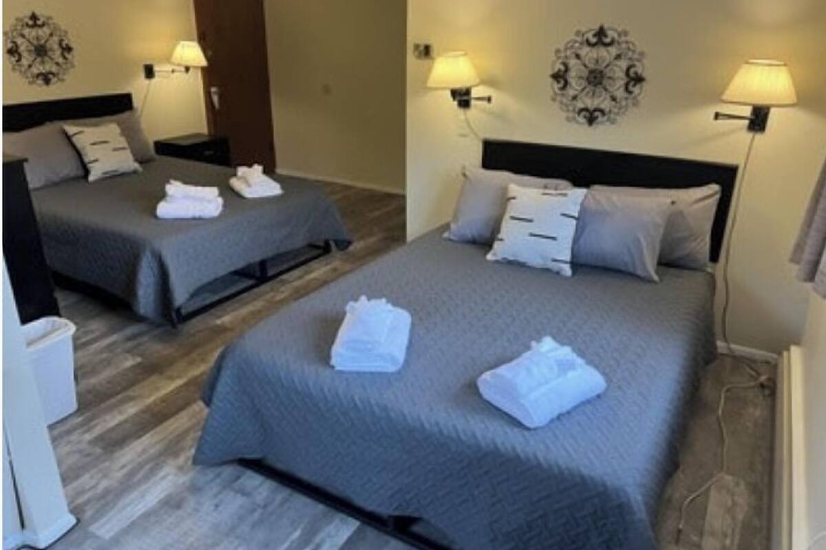 A hotel room with two beds, each with a grey bedspread and white towels. There are wall-mounted lamps on either side of the beds.