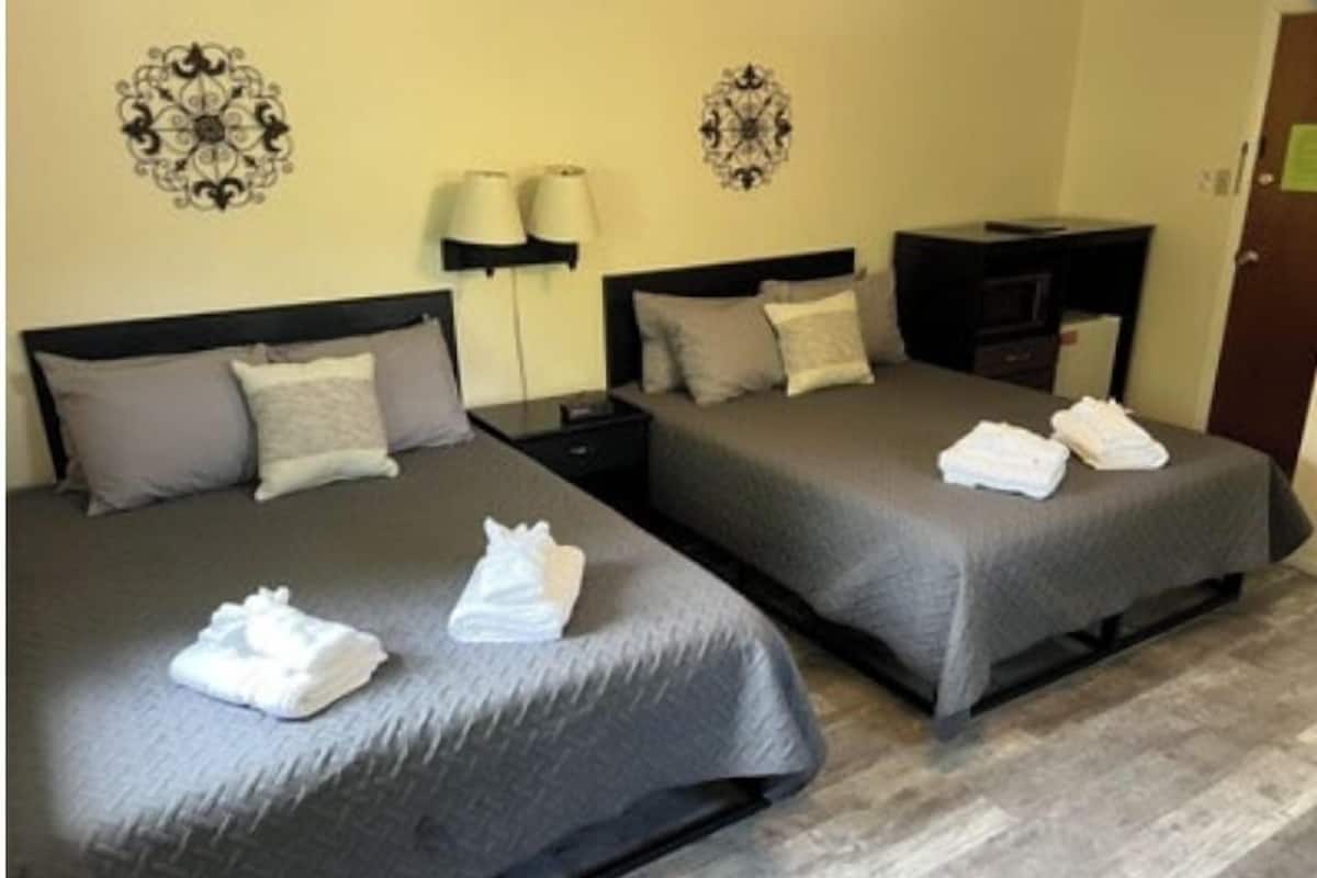A hotel room with two beds, each with a grey bedspread and pillows, a nightstand, a lamp, and decorative wall art.