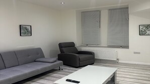 Ein modernes Wohnzimmer mit grauem Sofa, einem schwarzen Sessel und einem weißen Couchtisch. Zwei Fenster mit Jalousien, ein Heizkörper und eine kleine Pflanze auf der Fensterbank.